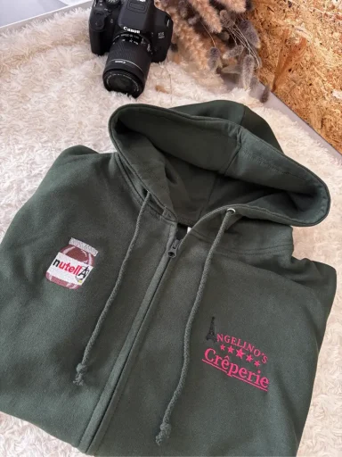 Grüne Kapuzenjacke mit gesticktem Logo und Kamera im Hintergrund.
