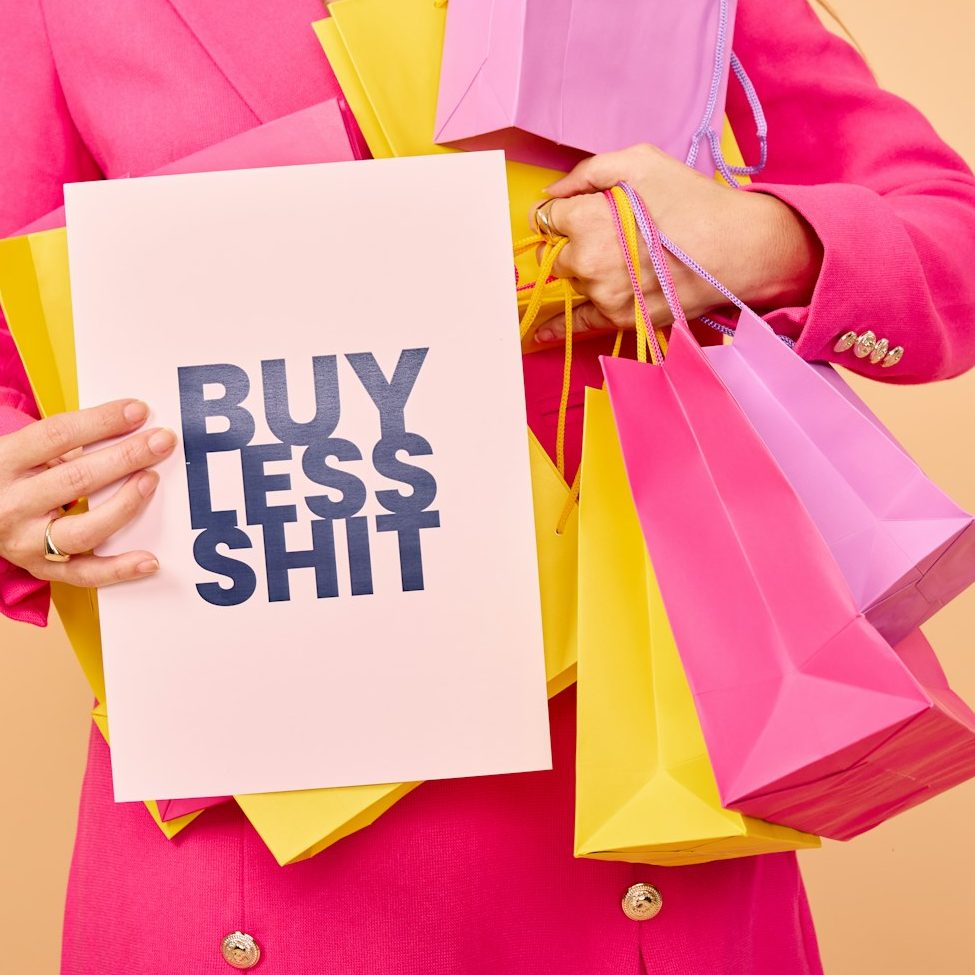 Person in pinker Kleidung hält ein Schild mit der Aufschrift „BUY LESS SHIT“ und Einkaufstüten.