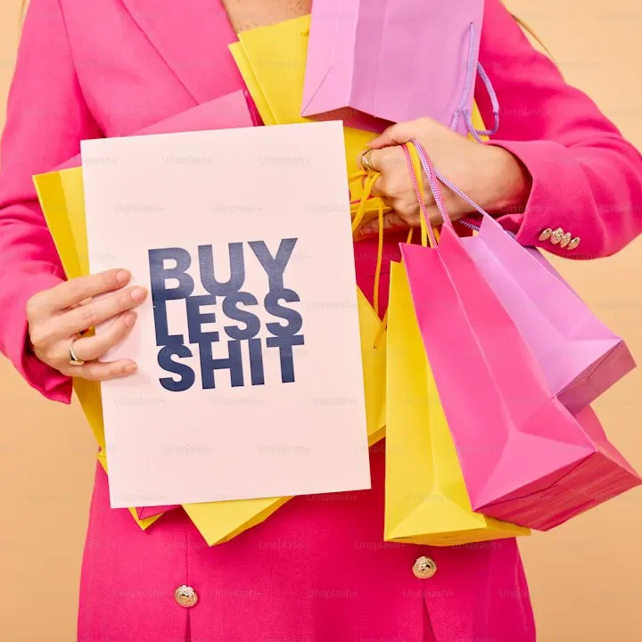 Person in pinker Kleidung hält ein Schild mit der Aufschrift „BUY LESS SHIT“ und Einkaufstüten.
