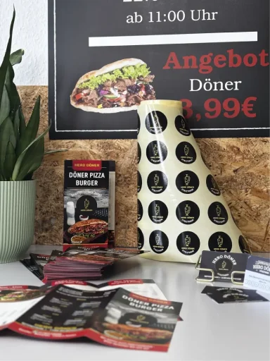 Döner-Spezialangebotstafel neben einem Topf mit Pflanzen und Werbeflyern auf einem Tisch.