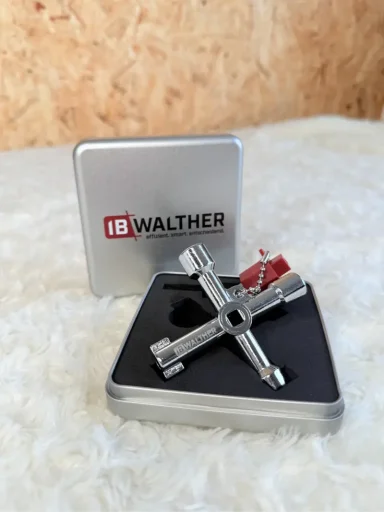 Ein silberner Schlüsselanhänger in Kreuzform, neben einer grauen Box mit der Aufschrift "IB WALTHER".
