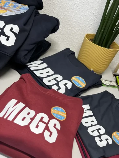 Stapel T-Shirts in Blau und Burgunder mit "MBGS"-Aufdruck und bunten Aufklebern.