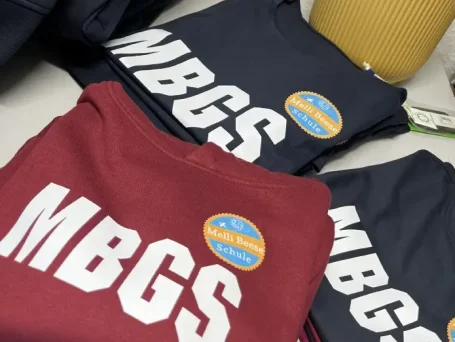 Drei T-Shirts in rot und blau mit der Aufschrift "MBGS" und einem Aufkleber.