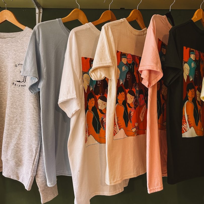 Sechs T-Shirts mit farbigen Grafiken hängen an Holzbügeln.