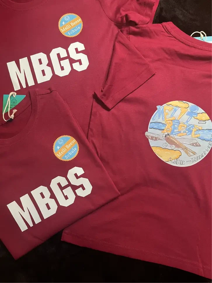 Rote T-Shirts mit dem Aufdruck "MBGS" und einem bunten runden Motiv auf der Rückseite.