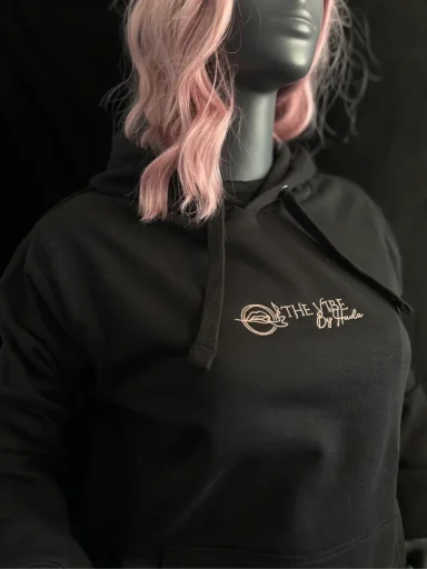 Schwarzer Hoodie mit goldfarbener Schrift und Kapuze, getragen von einer grauen Figur.