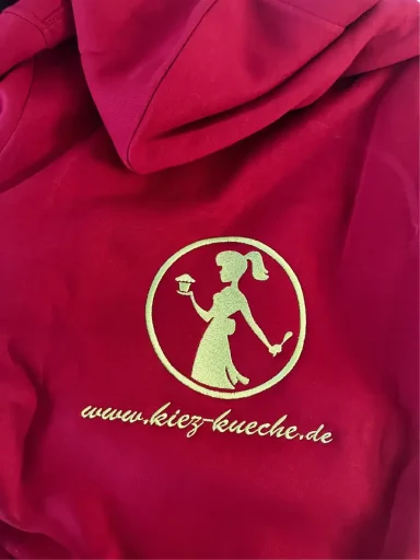 Rote Kapuzenjacke mit gelbem Logo einer stilisierten Frau und Website-Adresse.
