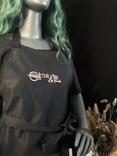 Schwarze Schürze mit Logostickerei, an einer grauen Figur mit grünen Haaren drapiert.