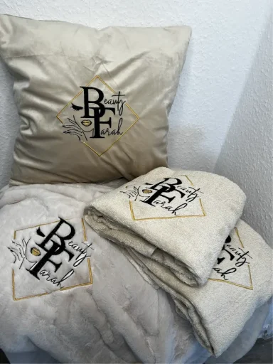 Beige Kissen und Decke mit gesticktem Logo in Schwarz und Gold.