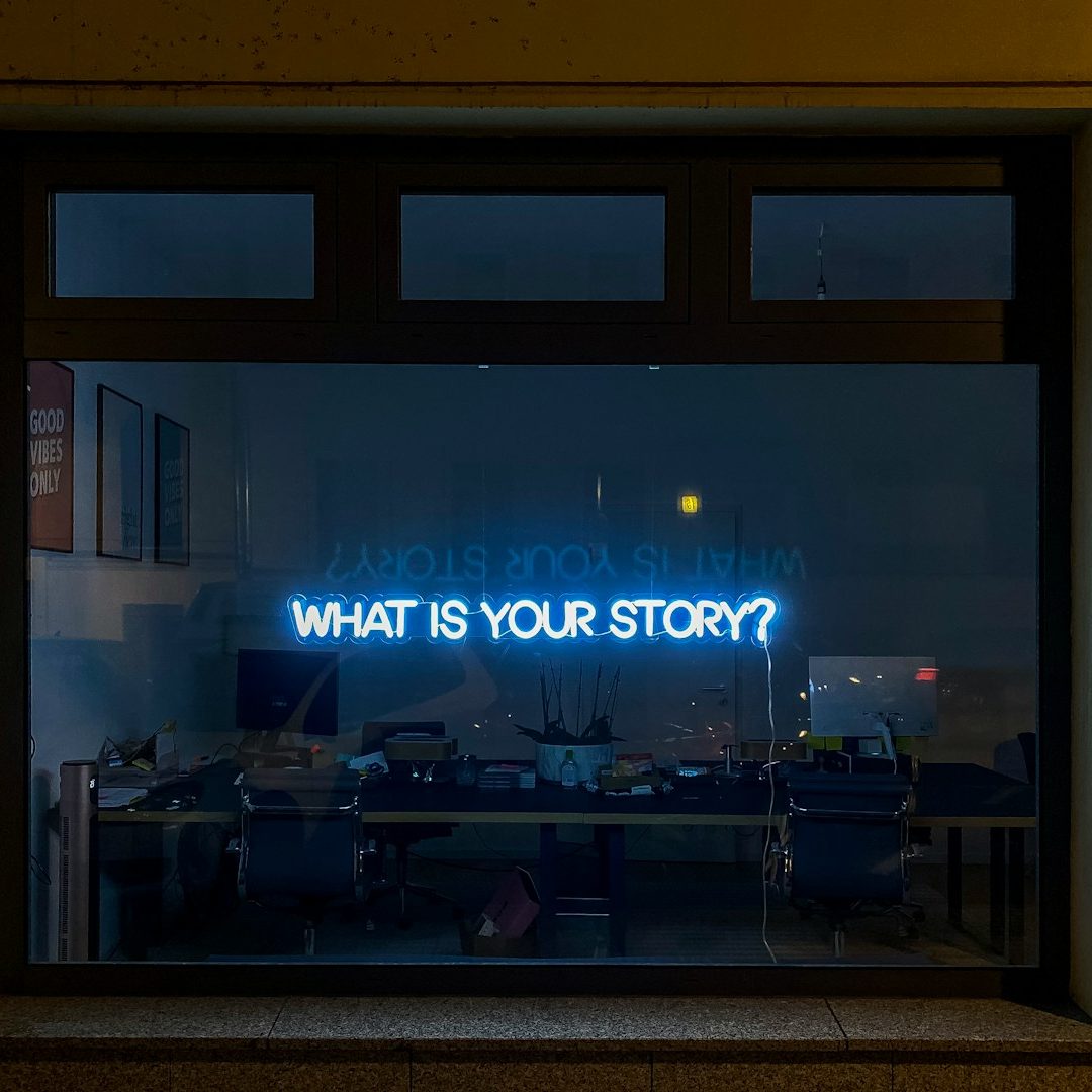 Neonlicht mit der Aufschrift "WHAT IS YOUR STORY?" in einem Fenster.