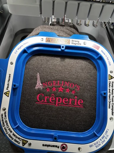 Text auf gesticktem Stoff: "Angelino's Crêperie" mit Eiffelturm-Symbol.