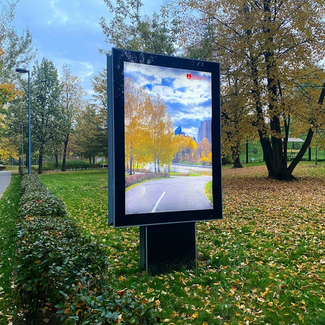 Werbetechnik für Unternehmen & Standorte Digital Werbetafel in einem Park mit herbstlichen Bäumen und einem Weg.