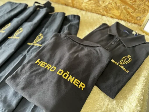 Schwarze Poloshirts mit goldenem Aufdruck "HERO DÖNER" auf hellem Hintergrund.