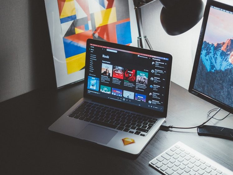 Laptop auf einem Tisch, daneben Monitor und weiße Tastatur, im Hintergrund Kunstwerke.