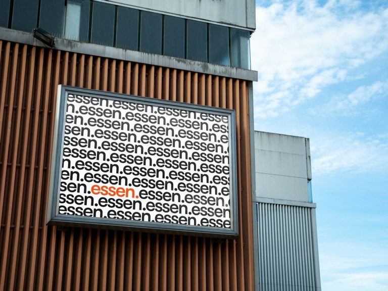 Ein Schild mit dem Text "essen" in vielfacher Ausführung in schwarz und ein Wort in rot, an einer modernen Gebäudeaußenwand.