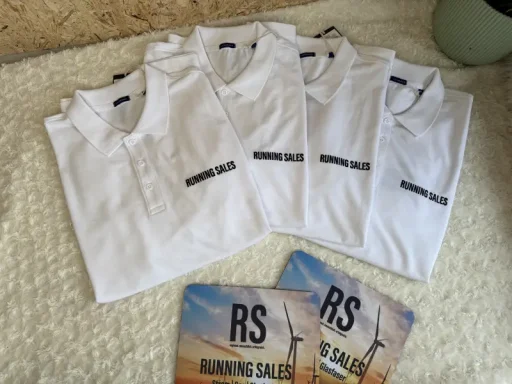 Vier weiße Poloshirts mit schwarzem Schriftzug und zwei Lizenzkarten darunter.