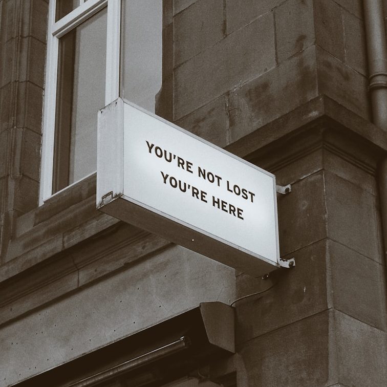 Weißes Schild an einer Gebäudeaußenwand mit der Aufschrift "You're not lost, you're here."
