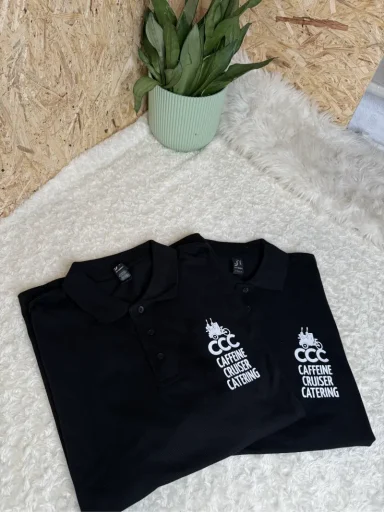 Zwei schwarze Poloshirts mit einem Logo, auf einer weißen Decke und einer grünen Pflanze.